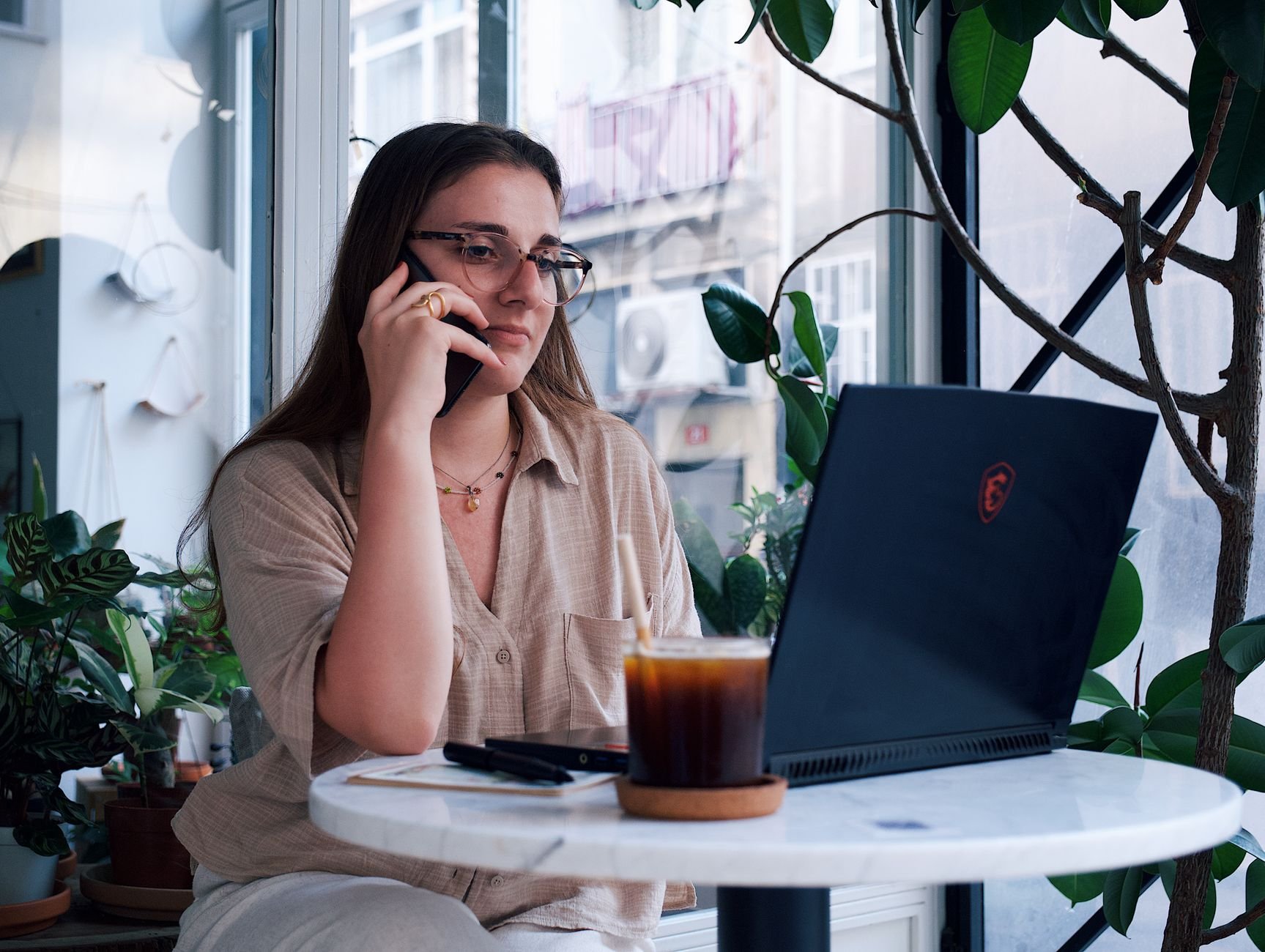 Young woman working on laptop and phone call in a cozy café with plants.